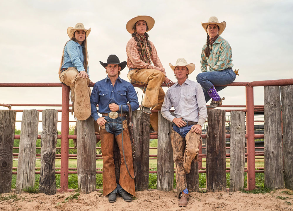 Three women and two men at a ranch wearing jeans, a shirt, cowboy hat, and cowboy boots.